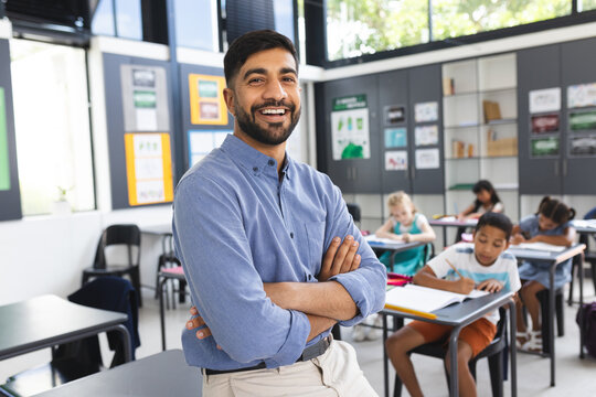 In school, Asian male teacher stands smiling with arms crossed in the classroom