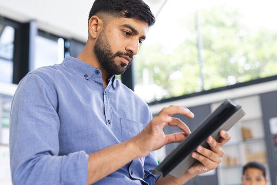 In school, Asian male teacher using tablet in classroom, a biracial male student watching