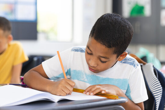 Biracial boy focused on writing in a notebook with a pencil in a school classroom