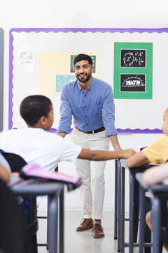 In school, Asian male teacher stands near board in classroom, young biracial boys sitting
