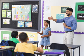 Young Asian male teacher teaching, biracial girl presenting in a school classroom