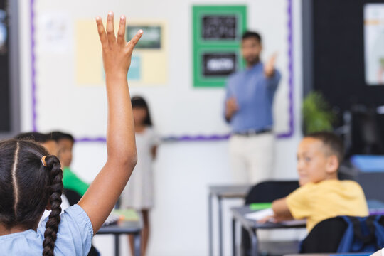 In school, young Asian male teacher stands by a board in the classroom, students raising hands