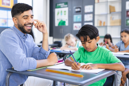A young Asian male teacher smiles, guiding biracial boy in writing