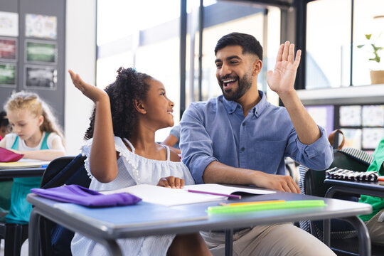 Young Asian male teacher and biracial girl high-five in a school classroom setting