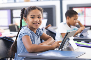 Biracial girl with braided hair smiles at the camera, using a tablet in a school classroom
