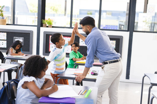 In school, young Asian male teacher giving high-five to a biracial male student in the classroom