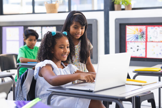 Two biracial girls, one in white dress, use a laptop in a classroom