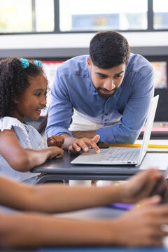 In school, Asian male teacher showing laptop to a biracial female student in classroom