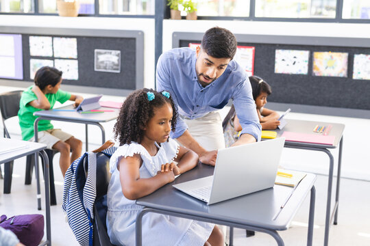 A young Asian male teacher helps biracial girl with her laptop