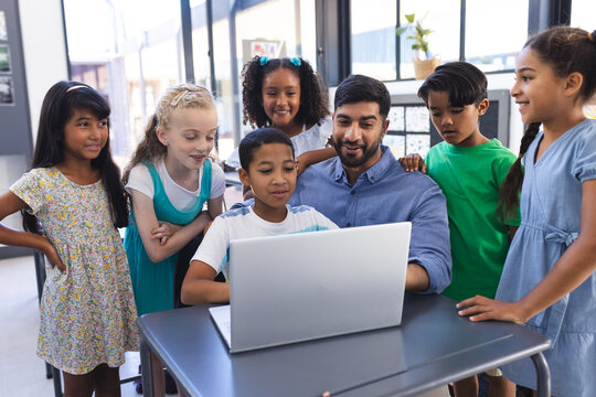 In school, young Asian male teacher and diverse students gather around a laptop in the classroom