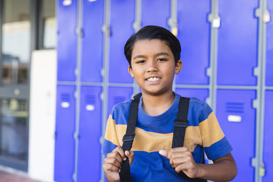 Biracial boy with a backpack stands in front of blue lockers in school