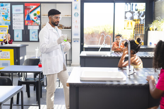 Asian male teacher with beaker stands by diverse children during science experi