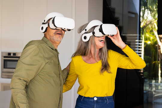 At home, diverse senior couple wearing virtual reality headsets in modern kitchen