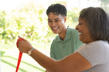 Young biracial male physiotherapist aids senior female patient at home with exercise band