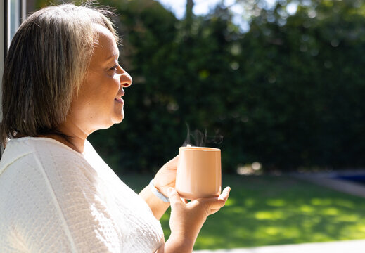A biracial senior woman holding steaming cup, smiling, looking outside, copy space - Powered by Adobe