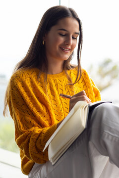At home, young Caucasian woman in yellow sweater writing in a notebook