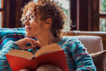 People in relaxation indoor leisure activity afternoon. One woman reading a book sitting comfortable on a chair and looking outside thinking and waiting. People enjoying relax time alone interior