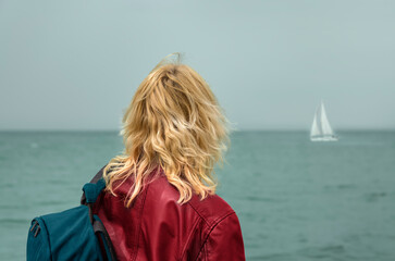 A woman in a red jacket and with a backpack on her shoulder looks at a sailboat at sea