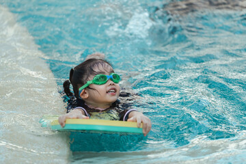 A young girl is swimming in a pool with a yellow and green board. She is wearing goggles and a...