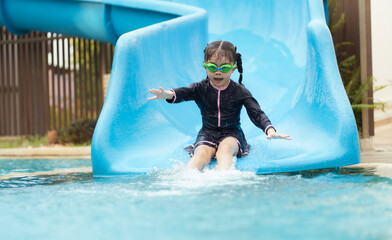 A young girl is sliding down a blue water slide. She is wearing a black swimsuit and goggles