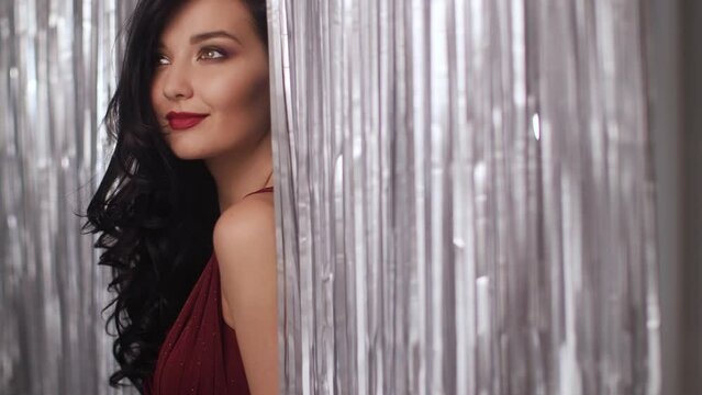 A glamorous beauty woman with long dark hair and red lipstick smiling behind a silver curtain, showcasing elegance and beauty.