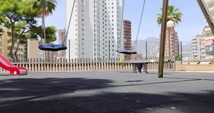 Footage of an empty children's playground showing the swings swinging on their own and a see saw in the park surrounded by high rise apartment blocks in the city of Benidorm in Spain