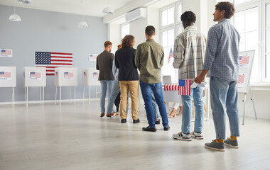 Large group of diverse people registering at polling station holding American flags in hands on election day. American voters standing in queue at vote center going to put ballots in voting booth.