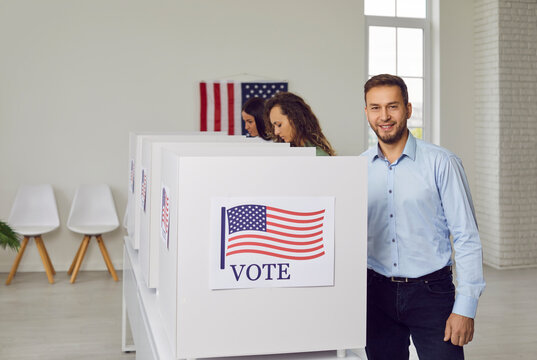 Group of American voters at ballot station during presidential elections in USA. Happy smiling business man in classic office shirt standing by white polling booth with US flag poster - Powered by Adobe