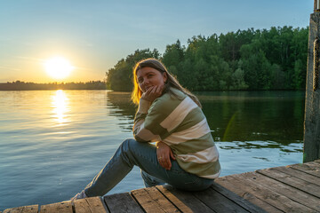 Adorable woman sits on a wooden dock, looking at the camera with a gentle smile. The sun is setting behind her, casting a warm glow over the lake