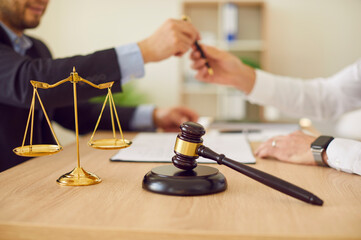 Close up of photo of male lawyer working on his workplace sighing a contract with a man client sitting in office at the desk with judge's gavel and scales in foreground. Legal law and advice concept.