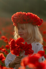 A young girl wearing a red flower crown is standing in a field of red poppies. She is holding a...