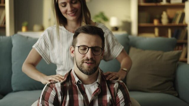 Caucasian man leaning back on his girlfriend sitting on couch in living room behind him, massaging neck and talking happily. Shot in slow-motion