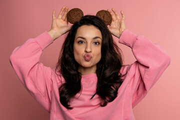 Funny dark-haired woman posing for the camera with oatmeal cookies on her head on the pink...