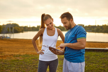 Young sporty man and woman watching the results of sports training outdoors checking time for performance in city park. Couple standing after doing sport jogging exercises in nature.