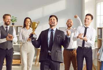 Businessman holds champion cup, celebrating success in office surrounded by colleagues who support him, applauding achievement. Highlights teamwork and joy of shared success.
