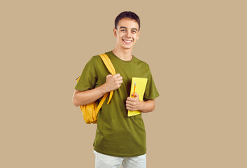 Portrait of happy high school or college student. Young man or teenage boy in casual clothes, with notebook and backpack standing isolated on beige color background, looking at camera and smiling