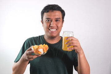 Fototapeta premium Enjoy healthy food. Handsome young asian man eating fruit salad and orange juice, smiling while standing on white background