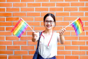 Closeup a cheerful Asian medical student action holding rainbow flag on brick block building wall...