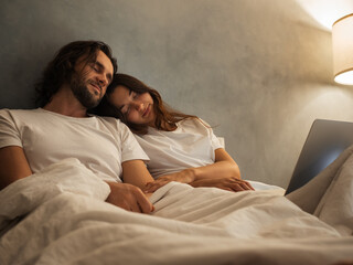 Peaceful couple sleeping in bed, resting against each other with a laptop nearby. Cozy nighttime...