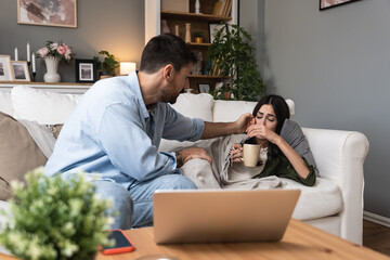 Young man helping and treats his sick woman at home who is wrapped in a blanket with a high body...