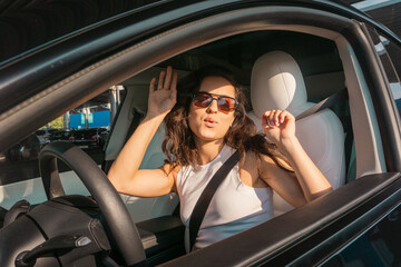 Smiling young woman in sunglasses driving vehicle on the road on a bright day while listening the...
