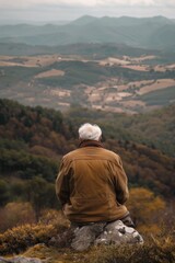 Naklejka premium Elderly man in a brown coat sitting on a rock, overlooking a vast and beautiful mountainous landscape.