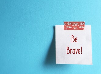 Stick note on blue copy space background with handwritten words BE BRAVE, to encourage to face and...