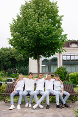 A group of young men sit on a bench in front of a large tree. They are all wearing white shirts and jeans. Scene is casual and friendly, as the boys are posing for a picture together