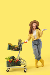 Female farmer with trolley of healthy food on yellow background