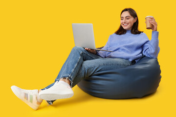 Pretty young woman with laptop sitting on bean bag chair against yellow background