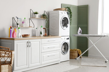 Interior of light bathroom with washing machines, houseplants and ironing board