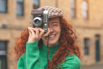 portrait of exited redhead woman taking pictures with vintage camera in city toned image