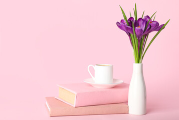 Vase with beautiful crocus flowers, cup of coffee and books on pink background