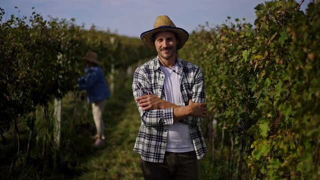 Young handsome farmer in hat standing in the vineyard, looking at camera and smiling. Two female winemakers ar standing in the background